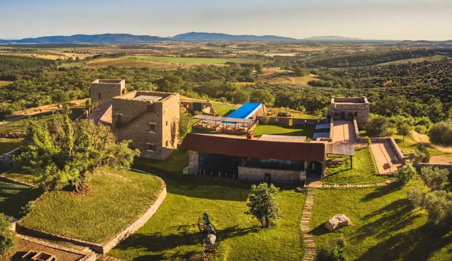 Aerial view of a restored historic stone estate with a pool, surrounded by olive groves and rolling countryside under warm sunlight